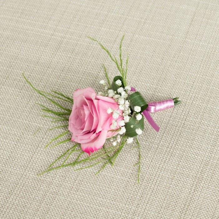 A finely detailed pink rose boutonniere is showcased against a pale beige, linen-style fabric, the gentle texture of the cloth echoing the understated elegance of a well-pressed suit ready for a Brent Cross ceremony. The single rose sits at the centre of the composition, positioned at a slight angle as if just set down by a careful florist. Its petals are velvety and fresh, starting with a soft, blush pink in the tightly nested middle and gradually shading to a richer, more saturated pink at the tips of the outer petals, where they curl open in a relaxed, romantic spiral. Gathered around the base of the rose are clusters of tiny white baby's breath flowers, each floret no larger than a pinhead, forming a soft halo that lightens the look and adds a traditional, almost vintage charm often chosen for North West London weddings. Behind the rose, fine, slender blades of green, grass-like foliage fan out in gentle arcs, creating a light, airy frame. A deeper green leaf, more substantial in shape, is neatly folded and wrapped around the stems, its edges clean and intentional, giving the boutonniere a tailored, structured finish. The stems are tightly bound in a smooth satin ribbon that shifts between lavender-pink and deeper magenta tones, secured in a simple, neat knot with short ends resting neatly on the fabric. Soft, diffused lighting emphasises the subtle shine of the ribbon, the matte weave of the linen, and the delicate contours of each petal. The overall mood is intimate and considered, ideal for a groom or special guest setting off from a Brent Cross home toward a registry office or local church, wearing a piece that feels both timeless and thoughtfully handcrafted.