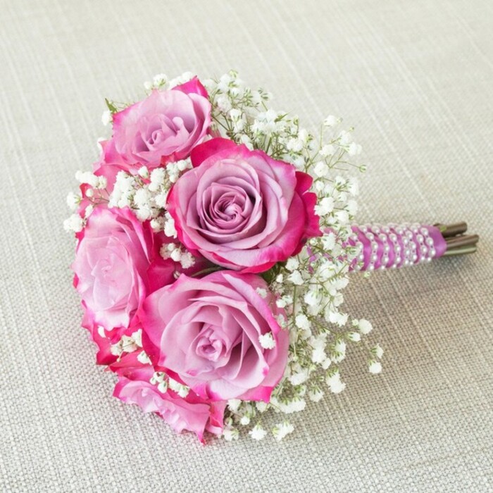 This sophisticated, compact bouquet showcases five radiant roses as its centerpiece, ideal for a wedding or special event in the Brent Cross area. Each bloom presents a striking transition from deep fuchsia at the outer petals to gentle lavender at the heart, producing a beautiful, multi-tonal gradient that speaks of tender romance and artistic workmanship. Interspersed between these lush, full roses are wispy stems of baby's breath, their tiny, white florets introducing volume and a delicate airiness to the overall structure. The bouquet's short, expertly bundled stem handle is dressed in a smooth purple ribbon, then graced with spiraling rows of dainty white pearls-a detail that feels at home in the classic yet modern vibe of events at nearby Brent Cross hotels or synagogues. Fresh green stems peek beneath the ribbon, confirming the bouquet's hand-picked freshness. The arrangement is pictured on an understated, oatmeal-hued linen reminiscent of north London's artisanal cafés, the subtle background allowing the bold colours and pearly details to shine. Photographed from above to highlight its rounded contours and careful styling, this elegant bouquet is perfectly suited for a bridesmaid, flower girl, or as a petite centrepiece, capturing the balance of classic beauty and local Brent Cross taste for refined, heartfelt celebrations.