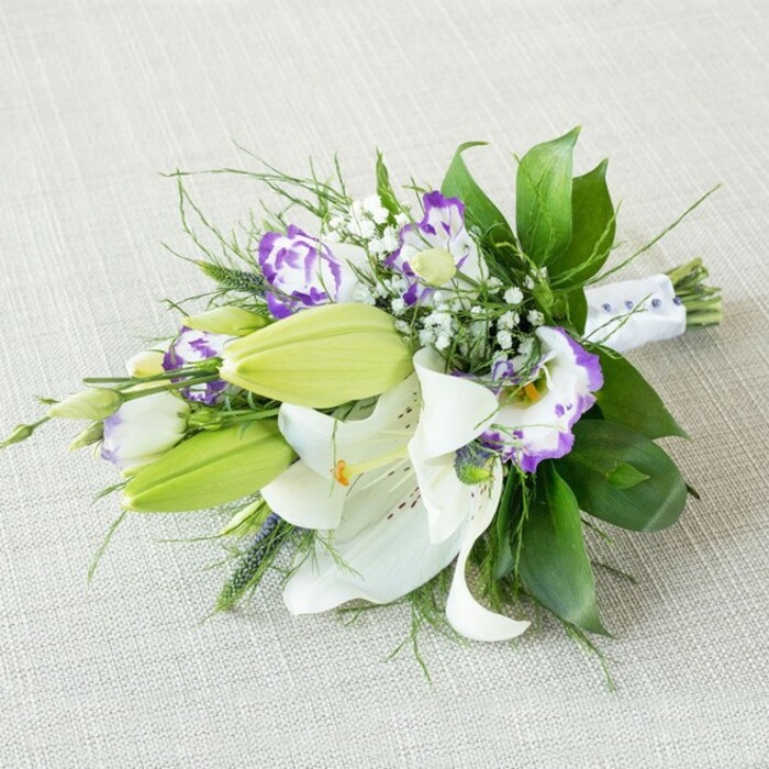 A refined, elongated bouquet of white lilies and purple lisianthus is photographed diagonally on a light beige, subtly woven fabric that recalls soft linen, the kind you might find on a carefully laid table in a Brent Cross family home. The stems are gathered at the right and fan outward to the left, giving the design a gentle sense of direction, as though ready to be lifted and carried down an aisle at a local ceremony. Dominating the arrangement are luxurious white Oriental lilies, some fully unfurled with broad, silky petals that curve away from rich yellow-orange stamens, lightly dusted with pollen and edged with subtle freckles of russet. Nestled among them are bright, lime-green buds, held upright and firm, providing contrast and the promise of new life and continuous blooming. Interlaced through the lilies run clusters of lisianthus, their blooms behaving like miniature, ruffled cups. Some petals blend creamy white with a blush of lavender, while others are more boldly rimmed in vivid purple, adding depth and a painterly quality to the palette. Wisps of baby's breath sprinkle the space between the main flowers like tiny white stars, softening the arrangement and adding a delicate, romantic haze. The greenery beneath and around the flowers is lush and varied, with sturdy, glossy leaves forming structure and finer, airy fronds extending lightly beyond the main outline, giving the bouquet a relaxed, garden-inspired feel that wouldn't look out of place in a park near Brent Cross. At the tied end, a smooth white ribbon, adorned with neat dotted beads or tiny pearls, cinches the stems into a comfortable grip, suggesting the work of a skilled florist who understands both beauty and practicality. The soft, balanced lighting ensures there are no harsh shadows, instead bringing forward every texture-the satiny lilies, the crimped lisianthus petals, the crisp greenery-making this bouquet ideal for illustrating sophisticated wedding flowers, graceful anniversary gifts, or elegant event décor available from a trusted local studio such as Brent Cross Flowers.