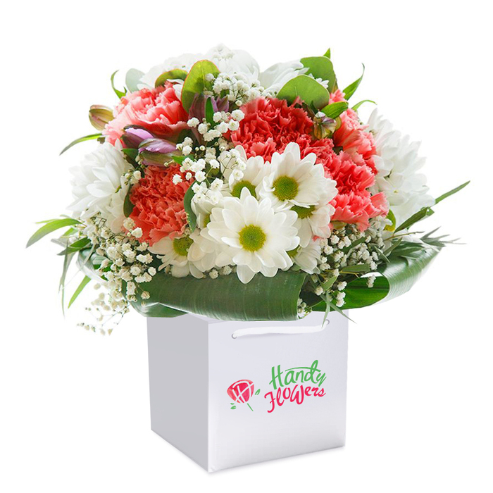 A masterfully arranged bouquet rises from a spotless, square white gift bag, its vivid colours and textures highlighted by a bright, seamless backdrop. This abundant arrangement intertwines soft coral, blush peach, and lively red carnations, each unfurling artful, ruffled petals that build softness and volume. Bright white daisy blooms adorn the bouquet, their faces aglow with fresh yellow-green hearts, reminiscent of cheerful displays at Brent Cross Shopping Centre. Baby's breath (gypsophila) threads its way throughout the composition, casting a fine, airy mist across the blooms and softening the overall feel. Peeking from one side, a handful of slender, purple-tinged buds bring layers and hints of soon-to-open promise. Anchoring the design, deep green, glossy foliage encircles the arrangement-a robust base that supports the bouquet's full, rounded form. Every element is gracefully balanced, a reflection of skilled, attentive floristry drawing upon years of local knowledge. The simple design of the white gift bag, finished with white ribbon handles, suggests the arrangement is ready for gracious giving-perfect for doorstep surprises from Brent Cross Flowers or thoughtful gestures to homes off Prince Charles Drive. The overall impression is one of generous celebration and enduring freshness, crafted with care for all the special moments that call for a touch of Brent Cross elegance.