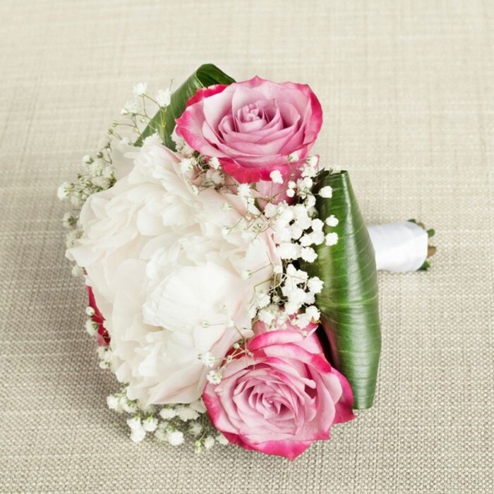Glowing under gentle, even light, this exquisite bouquet commands attention with its meticulous design-a true reflection of the floral artistry found in Brent Cross. Set upon a neutral, textured background, the arrangement brings together two striking pink roses whose petals gently transition from vivid fuchsia at the tips to soft, tender blush near their tightly coiled centers, symbolizing freshness and the quiet elegance of local events along Prince Charles Drive. Nestled at the heart lies a luxurious white peony, its countless ruffled petals layered in billowy, creamy folds, exuding both lushness and repose much like the tranquil atmosphere in Brent Reservoir's springtime. Dainty sprigs of baby's breath pepper the bouquet, their miniature white blooms dispersing a dreamy, cloud-like effect, filling the composition with weightless charm. Surrounding the flowers, deep green aspidistra leaves are expertly folded, offering a sleek, structured collar that draws attention to the vibrant interplay of pinks and whites while evoking the natural beauty of local green spaces. The bouquet's foundation is neatly secured with a clean white satin ribbon, providing not just stylistic flair but a comfortable grip-ideal for a bridesmaid preparing for a ceremony in the heart of Brent Cross or as a special delivery to a loved one nearby. Every element, lovingly selected and arranged by Brent Cross Flowers, is highlighted in the softly filtered light, making this creation a sophisticated statement piece for weddings, anniversaries, or heartfelt celebrations throughout the area.
