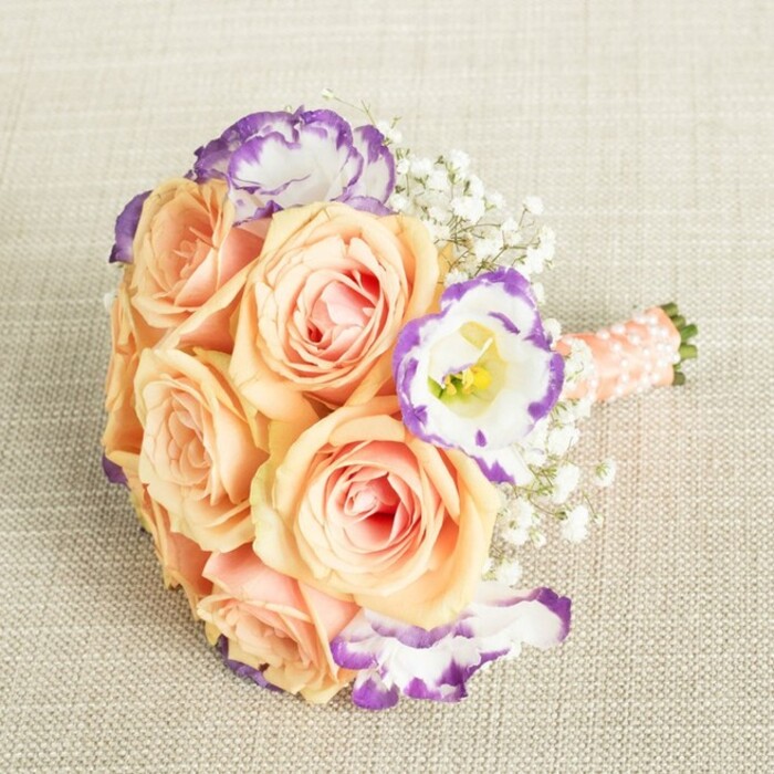 A romantic bouquet, poised on an oatmeal-toned linen, captures the spirit of celebration found in the heart of Brent Cross. At its core, generously blooming roses in apricot and soft peach are carefully placed, their swirling petals tinted with soft pink and ivory, exuding a fragrance that hints at fresh mornings along Brent Cross's leafy avenues. Punctuating this gentle backdrop, standout white lisianthus blossoms edged with striking purple lines add a modern artistic edge, balancing tradition with fresh local energy. Interlaced throughout, fine sprays of white baby's breath create whimsy and lightness, echoing the tranquil beauty found near Brent Reservoir. The bouquet's stems are elegantly gathered and enveloped in a smooth peach-pink satin ribbon, adorned with subtle pearl bead embellishments-a tactile, beautiful detail crafted with the precision familiar to Brent Cross Flowers. Ideal for weddings, bridal showers, or sophisticated events, this bouquet bridges classic floristry with contemporary flair, reflecting the unique blend of tradition and innovation growing in the Brent Cross community. The result is a harmonious arrangement, quietly radiating joy, refinement, and heartfelt connection.