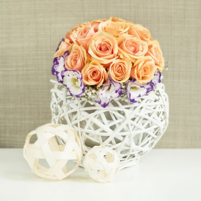 On a clean, white surface that reflects soft ambient light, an artfully composed table arrangement radiates gentle elegance, as if styled for a wedding table setting in a Brent Cross hotel or a chic dinner party along Hendon Way. At the centre of the design, a dense, spherical dome of peach and apricot roses rises proudly, each bloom luxuriously full and open. The roses are massed so closely that their velvety petals create an almost seamless, cushioned surface, with hues drifting from delicate blush to richer apricot and soft salmon, suggesting warmth, tenderness, and joyous celebration. Ringing the base of this rose dome is a band of white eustoma, or lisianthus, their frilled petals finely edged in deep purple or violet. This coloured trim adds a striking accent that frames the roses and gives the arrangement a refined, almost regal point of contrast. Delicate sprigs of white baby's breath are nestled between the lisianthus blossoms, contributing subtle texture and a light, airy quality that softens the overall form. The bouquet is securely positioned in a distinctive, broadly spherical container made from white woven wicker or rattan. Its open, lattice-like structure allows glimpses of the stems and interior shadows, lending the piece a sense of modern craftsmanship that fits well with contemporary Brent Cross interiors and newly renovated flats nearby. In the foreground, two decorative spheres fashioned from slim, pale wooden strips sit casually on the white surface, one larger and one smaller, echoing the container's open weave and extending the natural, tactile theme. Behind everything lies a neutral beige fabric backdrop with a gentle texture that keeps the focus on the rich interplay of colours and materials. The overall impression is of thoughtfully curated beauty, perfect for illustrating elegant wedding centrepieces, stylish home décor inspiration, or special occasion florals available in and around Brent Cross.