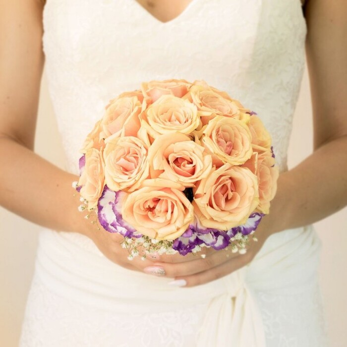 In this photograph, the camera lingers on the midsection of a bride, capturing her from just below the shoulders to the soft fall of her skirt, with the background rendered as a gentle blur of pale light. She is dressed in a classic white gown, its bodice overlaid with intricate lace that traces delicate patterns across the fabric, leading into a flattering V-shaped neckline. The waistline is accented by folds of smooth, slightly textured material that wrap elegantly around her middle before flowing downwards, suggesting movement even in stillness. Held carefully in both hands at the centre of the frame is a round bridal bouquet, crafted with the kind of precision you might expect from an experienced local florist serving weddings across Brent Cross and the nearby Golders Green area. The upper portion of the bouquet is a lush mound of roses in mellow peach, apricot, and blush-pink tones. Their petals are densely layered and gently curved, some blooms more open than others, creating a sense of depth and natural rhythm within the domed shape. Beneath this rose-filled crown sits a defined ring of deep purple flowers edged in creamy white, most likely lisianthus, whose ruffled petals lend a soft, frilled outline and a touch of drama. Threaded subtly among the stems near the base, sprigs of tiny white baby's breath form a mist-like border that lightens the silhouette and mirrors the lace on the gown. The bride's nails are neatly shaped and painted in a pale, understated shade with a whisper of sparkle on one thumb, adding a hint of personality to the otherwise classic look. The overall mood is one of quiet romance and poised anticipation, as if the bouquet has just been lifted for a final check before stepping out to a ceremony in a local Brent Cross church or hotel.