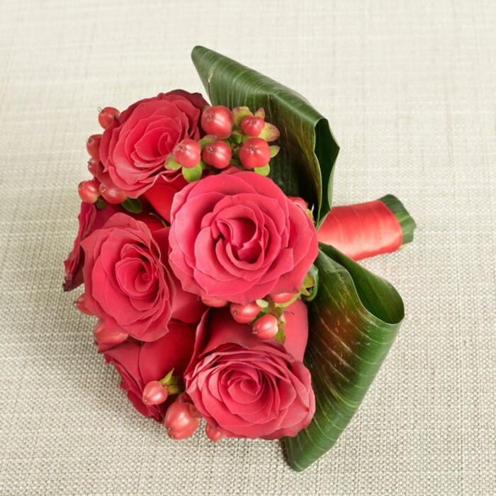 An intimate, close-up view reveals a beautifully composed bouquet of deep red roses resting on a soft, cream-beige fabric that has a faint, linen-like texture, similar to elegant tablecloths you might see in restaurants and hotels around Brent Cross. At the heart of the arrangement sit several large roses, each bloom at its peak, the petals dense and velvety, layering tightly in the centre and easing outward into gently curved edges that catch tiny traces of light. Their colour shifts subtly from almost wine-red in the shadows to a glowing crimson along the outermost petals, creating depth and a sense of movement within the fixed stillness of the photograph. Between the roses, clusters of small, rounded Hypericum berries in deep red and warm reddish-pink emerge on delicate stems, their smooth, polished surfaces contrasting against the plush softness of the petals and adding a festive, almost jewel-like sparkle. Around the lower portion of the bouquet, broad, dark green leaves have been carefully shaped and wrapped, folding in graceful arcs that both cradle and frame the central flowers, giving the composition a modern, structured edge while still feeling organic. A rich red satin ribbon gathers the stems tightly, visible to one side, its sheen echoing the luxurious mood of a special evening, perhaps a proposal in a quiet corner of nearby Hampstead or a milestone celebration at home in Brent Cross. The lighting is soft and diffuse, with no harsh glare, emphasising the natural richness of the reds and greens and highlighting the florist's craftsmanship. It feels like a bouquet chosen for anniversaries, romantic gestures, or Valentine's Day, composed with the careful eye of someone used to creating memorable moments for local couples and families.