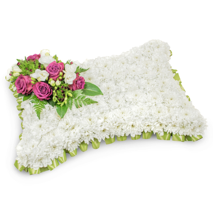 This rectangular memorial pillow is a quietly powerful expression of condolence, its entire face covered in a dense, uniform layer of small white chrysanthemums that create a soft, textile-like surface suggesting purity, calm and eternal rest. The chrysanthemums' compact petals and faint green stems combine to make a visually seamless white field, while a carefully placed accent on the top-left corner provides a point of focus and warmth. A cluster of deep magenta and fuchsia roses, rich and velvety, rises gently from the white bed; between them, delicate white freesias add lightness and a subtle, sweet fragrance that lifts the composition. Sprigs of green foliage - including slender fern fronds and smaller glossy leaves - cradle the colourful spray, giving it structure and a sense of living support. Encircling the base, a pleated trim of pale green satin ribbon peeks out from beneath the blooms, its soft sheen creating a neat, refined border that echoes the greens within the arrangement. Set against a pristine white background with a natural shadow to indicate dimension, the tribute's textures and hues read clearly: the pillow's plush white field, the roses' deep jewel tones, the freesias' creamy highlights and the fresh greens. Formally suited to funeral services, graveside placements or wake displays across Brent Cross and the surrounding neighbourhoods near the Brent Reservoir and Cricklewood Broadway, this design communicates rest, honour and a gentle, abiding remembrance.