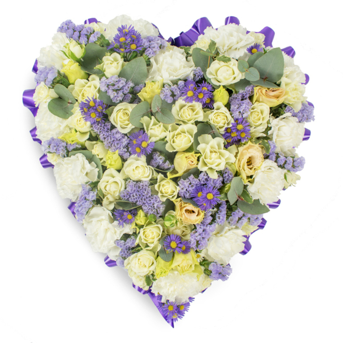 A lovingly arranged heart-shaped floral tribute stands out against a pristine white backdrop, its compact surface composed to convey comfort and reverence. Cream and pale yellow roses form the primary texture, their petals caught between tight bud and gentle bloom, some edged with a whisper of light green that creates a natural, living depth across the heart. White carnations fill the spaces between roses, giving the piece a soft, reassuring bulk and a traditional sympathy feel, while lavender-hued statice blankets small pockets with an airy, delicate texture. Bright purple, daisy-like asters with sunny yellow centres appear throughout, their saturated colour bringing occasional points of warmth and visual contrast amid the softer palette. The arrangement is neatly framed by a continuous, looped purple satin ribbon around the entire perimeter, folded to sit flat and define the heart's outline with a refined finish. Rounded, blue-green eucalyptus leaves are scattered thoughtfully to interrupt the floral mass with calming silvery tones and suggest the faint, cooling scent of fresh foliage. The studio lighting is even and gentle, allowing the creamy tones and pale lavenders to read as soothing rather than muted. This composition feels especially apt for funeral services, wakes or grave tributes-an expression of steadfast love, remembrance and consolation. Families in Brent Cross, particularly those visiting local churches or services near the shopping centre, often choose similar pieces for their dignified simplicity and comforting presence.