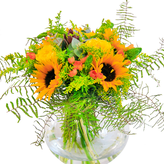 The first impression this bouquet gives is one of pure, sunlit exuberance-a mirror to Brent Cross's bustling, family-friendly atmosphere. Luminous sunflowers with generous golden petals are centered prominently, offering their warmth much like the sunlight streaming through windows along the North Circular. Surrounding them, plush yellow garden roses spiral open, their satin-soft petals unfolding amid a foundation of deep, glossy greens. Interwoven among these, clusters of bright red hypericum berries glimmer with a lacquered sheen that stands out beautifully against the matte yellows and leafy backdrop. Delicate fern fronds and sprigs of goldenrod arch gracefully, introducing airy movement reminiscent of a gentle breeze in Claremont Road's open spaces. Touches of silvered eucalyptus peek from within, adding complexity both visually and aromatically. Cradled in a perfectly round glass vase, the bouquet's stems are clearly visible, their lines echoing the clean, modern lines found in Brent Cross's renovated shopping centre. The fragrance is subtle but inviting, a blend of floral notes and fresh greenery that fills the air like the calm after a local rainstorm. This arrangement, designed for celebratory gatherings or as a spontaneous gesture of gratitude, illuminates any Brent Cross home-offering both beauty and the comforting sense of belonging that comes from a bouquet made by florists with deep ties to the NW4 community.