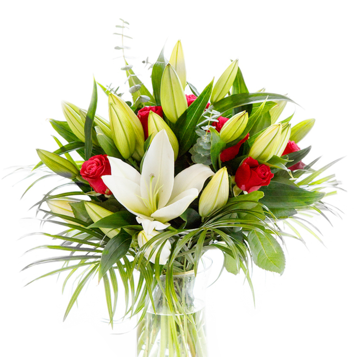A single open creamy-white oriental lily anchors this striking bouquet, its satin-smooth petals slightly cupped to expose pale yellow stamens and a pale green pistil that draw the eye inward. Surrounding it are numerous lily buds in lime-green and soft yellow hues, some tight and expectant, others beginning to unfurl - the arrangement suggests slow motion, a promise of more blooms to come. Interspersed among the lilies are clusterings of deep red rosebuds and small roses, their velvety petals still curled, offering concentrated notes of passion and classic romance against the lily's serene palette. The greens have been chosen for contrast and gesture: glossy dark green lily leaves create structure; long, arching palm-like fronds in a fresher emerald hue cascade outward with graceful rhythm; and rounded, silvery eucalyptus stems soften the composition with cool, muted tones. The flowers sit in a straightforward, cylindrical clear glass vase so the light-green stems and clear water are visible, lending the arrangement a clean, honest look. Shot on a bright white background, every texture and shade is emphasised - the skin of petals, the sheen of foliage, the translucence of water - producing a fresh, celebratory image. This bouquet, crafted by Brent Cross Flowers in our local shop, reflects neighbourhood sensibilities: it would equally suit a quiet anniversary in a Hendon flat or a cheerful congratulations dropped off near Brent Cross Shopping Centre, communicating care, purity and heartfelt intent without fuss.