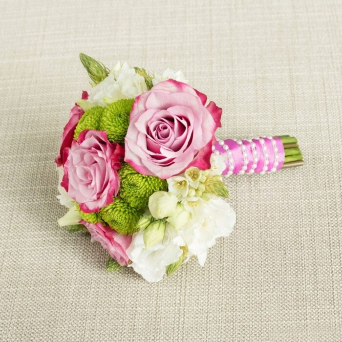 Laid horizontally on a softly textured, light tan fabric that could be a linen or burlap table runner, this petite bouquet looks as if it has just been set down between photographs at a Brent Cross wedding or engagement gathering. The arrangement centres around three lush pink roses, two of them fully open and one slightly more nestled, their petals layered like fine silk. Each rose carries a gentle gradient of colour: a tender blush pink at the inner whorls that gradually intensifies to a more saturated magenta-pink along the outer edges, giving a sense of depth and craftsmanship that feels very much like the work of an experienced local florist. Tucked intimately between the roses are rounded clusters of lime-green button chrysanthemums, their vividly coloured, tightly packed florets forming small, spherical cushions. These mums add a crisp, almost zesty freshness to the palette, balancing the romance of the roses with a playful, contemporary accent. Scattered through the bouquet are soft white and cream blossoms-likely lisianthus or freesia-whose slightly flared, multi-petalled blooms appear airy and delicate, adding lightness and a whisper of fragrance one might imagine catching while walking past flower stalls on the way to Brent Cross Shopping Centre. Tiny starry white flowers with golden centres, reminiscent of waxflower, peep through the main blooms, contributing fine texture and a subtly rustic charm. The stems are cleanly trimmed and gathered, then wrapped in a rich fuchsia satin ribbon that glows gently under the even, natural-style lighting. Over this ribbon, a single strand of petite white faux pearls has been wound in diagonal bands, crossing like a delicate lattice and finishing in an elegant, polished handle. The result is a compact bouquet that feels ideal for a bridesmaid, a younger bride, or a chic table arrangement for a small celebration at home in Brent Cross, radiating romance, freshness, and thoughtful detailing in every element.