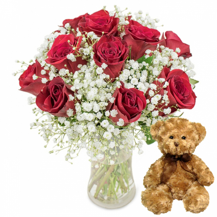 A beautifully balanced still-life arrangement is presented against a bright white background, highlighting every curve and colour in this romantic gift set. On the left, a tall, straight-sided glass vase holds a cluster of green rose stems, which can be clearly seen through the transparent surface and the softly glinting water inside. Above the vase, forming a rounded, generous dome of colour, are around twelve red roses arranged with expert precision. Each rose displays velvety petals in a sumptuous, deep crimson shade, the tones subtly shifting from brighter red at the outer edges to darker, wine-red shadows toward the centre. Some blooms are fully open, showcasing their layered spirals, while others are just beginning to unfurl, suggesting a bouquet with several days of beauty ahead. Winding gently throughout the roses are sprays of white baby's breath, their minuscule blossoms clustered together to create a cloud-like frame of delicate white around the richer red. This contrast of colour and texture is a favourite choice among customers ordering romantic flowers for delivery in Brent Cross, whether for anniversaries, proposals, or surprise celebrations after a day of shopping nearby. Touches of dark green foliage peek between the flowers, grounding the composition and adding a natural depth. To the right of the vase, on the same white surface, sits a medium-sized teddy bear made from soft light brown plush. Its fur looks fluffy and warm, with slightly rounded limbs positioned in a relaxed seated pose. The bear's face is sweet and inviting, with small black button eyes, a petite embroidered nose, and a faint stitched smile that together convey friendliness and comfort. A dark brown satin ribbon tied neatly around its neck introduces a subtle note of elegance, its sheen contrasting gently with the bear's matte fur. The overall mood of the image is intimate and heartfelt-a classic red rose and teddy bear combination that feels perfectly suited to Valentine's Day, milestones, or romantic gestures shared across the Brent Cross community.