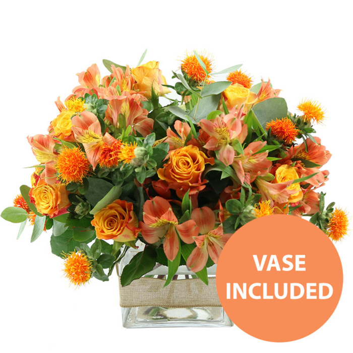 This image presents a generous, autumn-inspired bouquet of orange and yellow flowers, photographed in bright, even light against a stark white background so that every detail stands out. Dominating the centre are several large orange roses, their petals swirling outward in rich, layered spirals that transition subtly to a lighter, almost golden yellow near the centre in some blooms. Nestled between them are vivid yellow roses and garden-style blooms, their tighter, more compact petals contributing a slightly more structured look. Weaving through the design are stems of alstroemeria, the Peruvian lilies dressed in shades of peach, apricot, and soft orange. Their slender petals open wide, showcasing delicate streaks and speckles of deep rust, brown, and saffron inside each throat, as though each flower had been hand-painted. Small, spherical orange blooms with a distinctly spiky surface-resembling safflower or thistle heads-are dotted throughout, adding a playful, textural twist that contrasts with the softness of the roses. Surrounding this warm heart of colour is an abundance of foliage: elegant eucalyptus leaves in a dusty, silvery green extend outward, accompanied by richer, darker green leaves that anchor the arrangement, similar to the layered greenery glimpsed in gardens around Brent Cross and Golders Green. The bouquet is carefully arranged in a clear, square glass vase, its sides revealing the neatly cut stems and the clarity of the water, emphasising freshness and craftsmanship from the local florists at Brent Cross Flowers. A band of natural beige burlap or jute is wrapped around the vase's midsection, adding a rustic, handmade charm that softens the contemporary glass. In the image's bottom right corner, a bold, circular orange graphic with crisp white lettering reads VASE INCLUDED, signalling that the arrangement arrives as a complete, display-ready gift. The overall feel is warm, celebratory, and welcoming, ideal for birthdays, anniversaries, or bringing a cosy burst of colour into a home near Brent Cross.