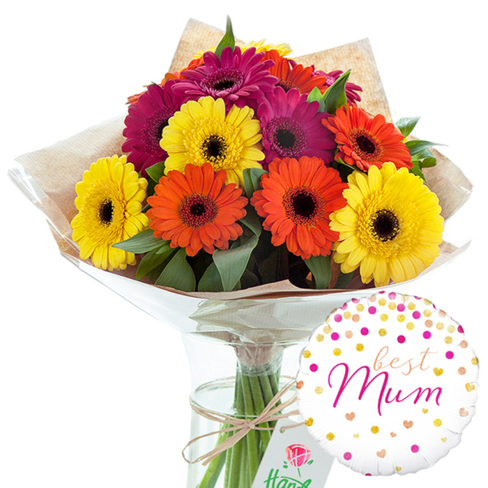 This image showcases a complete floral gift ensemble, the kind of thoughtfully styled gerbera bouquet and balloon pairing you might choose when sending flowers across Brent Cross and the nearby residential streets. The central focus is a voluminous bouquet of gerbera daisies, gathered into a rounded, almost domed shape. Each bloom is large and well-formed, with flat, evenly spaced petals radiating from a dark, textured central disk. The colour palette is distinctly warm: vibrant yellow gerberas add light, while zesty orange and deep magenta or fuchsia flowers provide depth and richness. Interspersed between the blooms are clusters of dark green leaves, which create a layered, natural backdrop and emphasize the brightness of the petals, reminiscent of flower beds seen in local parks on a sunny morning. The stems are carefully hand-tied and wrapped in a layer of rustic brown kraft paper, visible in soft folds around the base of the bouquet. This is enclosed in an outer shell of clear, glossy cellophane that hugs the flowers and catches reflections, giving the arrangement a neat, professional finish. Around the gathered stems, a simple length of natural twine is tied into a small bow, reinforcing the honest, handmade charm of the design. The bouquet stands upright in a tall, straight-sided glass vase filled with clear water, where the trim green stems can be seen in parallel lines. Just to the lower right of the vase, a single round foil balloon conveys the theme of the gift. The balloon's white background is peppered with metallic gold and shades of pink confetti-style dots and tiny heart shapes. At its centre, the words best Mum appear in elegant pink script, clearly legible. The overall composition feels warm and affectionate, a visual expression of appreciation perfect for Mother's Day, a milestone birthday, or an everyday thank-you to a mother figure in Brent Cross.
