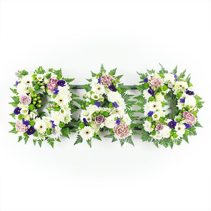 Three floral letters, carefully arranged in a straight horizontal line, spell out DAD against a clear white background, allowing the design to feel serene and uncluttered. Each letter is constructed as a solid panel of white flowers, primarily tight-headed chrysanthemums and daisy-like gerbera blooms, whose layered petals form a thick, cushiony surface that looks both soft to the touch and visually substantial. The subtle differences in petal size and the slight creamy nuances within the whites give the arrangement a gentle depth rather than a flat, uniform look. Around the edges of each letter, fresh greenery forms a delicate border: mossy-toned fern fronds and other fine foliage extend just beyond the flower edge, outlining the shapes and lending them a natural, garden-inspired character familiar to anyone who walks through the green pockets near Brent Cross Shopping Centre or along nearby residential streets. Woven through the white base are carefully placed accents of colour, never overpowering but thoughtfully positioned: rich violet and deep purple flowers, possibly spray chrysanthemums or small carnations, add a hint of formality and poise, while soft lavender roses and pale mauve blooms bring a comforting, almost nostalgic warmth that feels fitting for a memorial. The lighting is soft yet clear, ensuring the whites remain bright and the greens look fresh, as though the tribute has been prepared with newly delivered flowers ready for a same-day funeral service in Brent Cross or surrounding North West London. Overall, the DAD tribute reads as a graceful and deeply personal expression of gratitude and farewell to a much-loved father, created with sensitivity and professional skill.