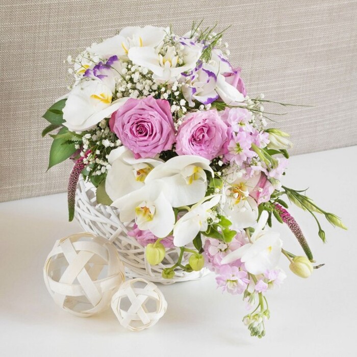 A graceful floral arrangement in a soft pink and white palette sits proudly in a round white wicker basket, as though placed at the centre of a wedding table in Brent Cross. The main focus is on generously open pastel pink roses, their petals unfolding in flawless spirals from tight centres to full, velvety blooms that appear almost touchable, suggesting tenderness and devotion. Interlaced with them are pristine white Phalaenopsis orchids, each blossom wide and smooth, their curving petals framing golden yellow centres flecked with subtle markings, adding a sleek, modern elegance to the design. Between these signature flowers, white Lisianthus blooms introduce a layered, ruffled texture; some display a faint brush of lavender-purple along the petal edges, lending a cool, sophisticated nuance to the palette. Slender stems of blush-pink stock rise from within the arrangement and drift outward, their clusters of small flowers forming airy plumes that bring gentle fragrance and a sense of relaxed, garden-inspired abundance, reminiscent of blooms gathered near Clitterhouse Playing Fields. Delicate sprays of baby's breath are woven throughout, tiny white flowers catching the light like a soft haze and connecting each element with a fine, ethereal texture. Fresh green foliage anchors the arrangement, peeking between blossoms and trailing subtly over the basket's rim, hinting at new buds and growth. The tightly woven white wicker basket rests on a smooth white surface that suggests a formally laid table or bright home interior, accompanied by two decorative orbs formed from pale, interlaced strands of natural material that mirror the basket's weave and introduce playful, organic shapes. Behind it, a soft beige background with a linen-like texture offers warm neutrality and allows the colours to stand out clearly. The scene evokes the atmosphere of a refined celebration, the sort of thoughtfully crafted centerpiece Brent Cross Flowers might deliver for weddings, anniversaries, or intimate family dinners.