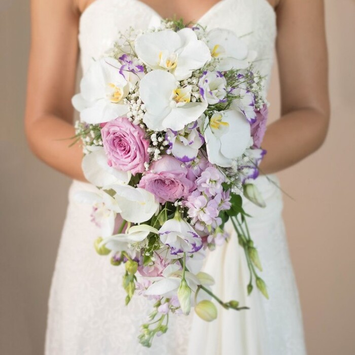 The image reveals a Brent Cross bride, seen from her chest to waist, gracefully holding a showpiece cascading bouquet before a subtly blurred neutral background, reminiscent of the soft light that fills local chapels on wedding mornings. The flowers are arranged in a luxurious teardrop silhouette, crafted by the expert hands at Brent Cross Flowers. Pristine white phalaenopsis orchids-at their peak, with wide, open petals and gentle yellow accents-anchor the composition, infusing the bouquet with elegance and drama. These are flanked by full, blush-pink garden roses, whose velvety, layered petals add classic romance and depth. Wisps of airy lavender and pale purple stock flowers thread vertically, complemented by delicate lisianthus-some edges tinged in deep purple-that introduce subtle movement. Intermingled throughout, frothy white baby's breath and hints of green foliage lift and lighten the arrangement, preventing the blooms from overwhelming the eye. The bride's white strapless gown, with lace detailing on the bodice and skirt, peeks into the frame, its intricate patterns echoing the natural intricacies of the flowers she holds. The bouquet cascades in a sweeping, natural flow, inviting thoughts of outdoor celebrations in Brent Cross gardens and creating a resonant sense of joyful anticipation perfect for local wedding inspiration.