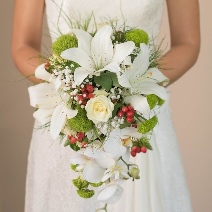 A striking, close-up view captures a Brent Cross bride holding a gracefully cascading wedding bouquet against the delicate lacework of her ivory gown, her presence exuding poised anticipation on her special day. The bouquet's design revolves around large, white oriental lilies, their sweeping petals touched by understated speckles, radiating chic sophistication. At its core, a perfectly formed cream rose draws the eye, nestled among sprightly lime-green cushion mums that lend fresh, contemporary contrast-reminiscent of springtime along Claremont Road. Wisps of silvery-white baby's breath fill the spaces between, creating a soft, cloudlike frame for the showier blooms. The bouquet's captivating silhouette is defined by an artful cascade of white phalaenopsis orchids, their long, elegant sprays descending gracefully and bringing a sense of fluid motion. Scattered through the display, vibrant red hypericum berries inject joyful color and a dash of North London exuberance. Outlining the arrangement, tufts of grass-like greenery and glossy foliage add further dimension, their varied textures reflecting a local taste for botanical diversity. The bouquet is masterfully balanced-lush yet refined, modern yet rooted in timeless wedding tradition. Set against the subtle texture of the bride's lace dress, this arrangement becomes a symbol of the union between heritage and fresh beginnings, echoing the warmth and stylish sensibility of Brent Cross weddings.