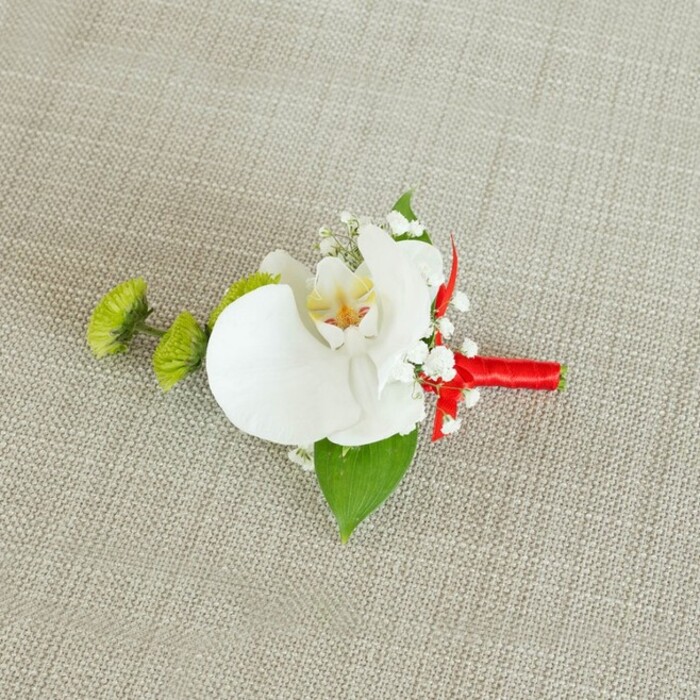 A sophisticated floral boutonniere lies against a pale beige, linen-like fabric, its subtly coarse weave creating a warm, natural canvas that lets the flowers shine without distraction. At the centre of this delicate arrangement is a single white phalaenopsis orchid, its wide, satiny petals opening in an elegant arc that feels both modern and timeless. The throat of the orchid reveals soft yellow tones gently melting into fine reddish-orange and light brown details, a painterly pattern that adds intrigue to the otherwise calm white bloom. Surrounding the orchid is a halo of white baby's breath, thousands of tiny blossoms clustered together like a light, misty veil, softening the overall look and lending a tender, romantic feel. To the left, two rounded lime-green chrysanthemum buds, tightly packed and slightly spiky in texture, bring a bright jolt of colour and a contemporary edge that would suit stylish celebrations around Brent Cross, from hotel receptions to intimate garden ceremonies. A broad, rich green leaf sits beneath the orchid, acting almost like a natural frame, while a smaller, darker leaf peeks out near the base, adding depth and contrast. The stems of the flowers are neatly gathered and wrapped with a vibrant crimson red satin ribbon, the wrap smooth and precise, ending in a small, neat bow that introduces drama and passion in a single stroke of colour. Gentle, even lighting highlights the sheen of the orchid petals and the delicate structure of each baby's breath flower, suggesting the careful hands of a skilled florist who understands both texture and balance. This boutonniere feels tailor-made for a groom or groomsman walking through the streets around Brent Cross towards a day filled with vows, photographs, and quiet, unforgettable moments.