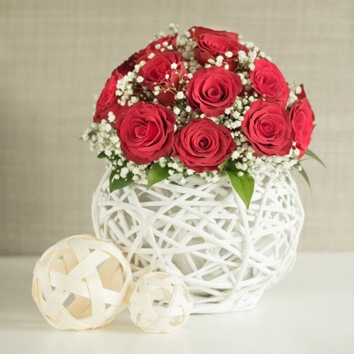 A thoughtfully styled floral arrangement of red roses and baby's breath sits on a pristine white table, evoking the atmosphere of a carefully dressed wedding reception near Brent Cross station. The focal point is a rounded bouquet of roughly twelve to fifteen roses, all in a deep, saturated red. Each rose is fully bloomed yet still firm, its petals layered tightly into a luxurious spiral that catches the light on every curved edge. Interlaced among these crimson flowers are generous sprays of white baby's breath, their clusters of tiny blossoms forming a delicate mist that lightens the whole design. A few hints of dark green foliage peep out from beneath the blooms, adding a discreet touch of natural contrast. This lush crown of flowers rises from a distinctive, bulbous white vase with an open, woven structure, as though slender white rattan or wicker strands have been wrapped and interwoven to create a hollow sphere. The open lattice allows glimpses of the stems within and gives the piece an airy, contemporary look that would suit modern homes off Hendon Way or around the Brent Cross shopping area. In front of the vase, resting casually on the same smooth white surface, are two decorative balls crafted from pale, flat strips of material twisted into open orbs. The larger one sits slightly closer to the viewer, while the smaller nestles just behind it, their creamy hues echoing the vase without overshadowing the flowers. Behind, a softly patterned, neutral wall in light beige or off-white forms a peaceful backdrop, gently blurred so that all attention falls on the roses' vivid colour and the interplay of woven textures. The scene communicates romance, sophistication, and the thoughtful touch of an experienced local florist from Brent Cross Flowers.