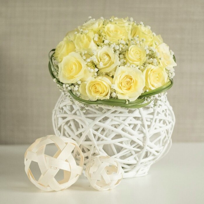 A beautifully composed wedding-style centerpiece sits in still focus, formed from a generous cluster of pale yellow roses arranged into a smooth, dome-shaped bouquet. The roses are tightly grouped, with each flower fully open so that the petals overlap and create a plush, textured surface, reminiscent of soft buttercream on a carefully iced cake. Threaded between the roses, airy white baby's breath punctuates the yellow with tiny bright dots, lending the arrangement a sense of lightness that feels particularly suited to spring and summer celebrations in Brent Cross. Around the base of the bouquet, a slender band of glossy green foliage is wrapped with great precision, acting as a natural ribbon that frames the flowers and offers a sharp, fresh transition into the container. The vase itself is a striking piece: white, woven, and sculptural, made from crisscrossing strands that form an open basket-like shape, slightly cinched at the middle. The open weave creates glimpses of the interior and allows the surrounding light to filter through, leaving intricate shadows on the white tabletop beneath. To one side of the vase, two decorative balls made from light, natural materials lie in relaxed balance, their open, geometric strands echoing the vase's texture and tying the whole vignette together, much like coordinated table décor at a reception venue close to Brent Cross Shopping Centre. The background is a soft, neutral beige, out of focus and textural like fine fabric, providing a calm, elegant setting that allows the creamy yellows, pure whites, and fresh greens to stand out. The image conveys an atmosphere of polished simplicity and gentle joy, perfectly at home in a wedding breakfast, anniversary celebration, or stylish dinner party in the local area.