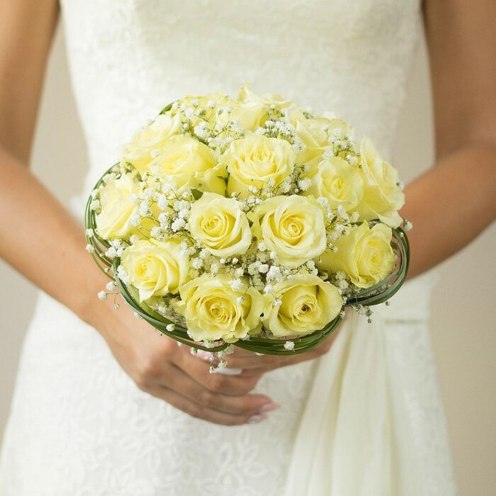 A refined, close-up image captures a moment brimming with the quiet joy and anticipation of wedding day preparations in Brent Cross. The bride, shown from the shoulders down in a luminous white gown, delicately cups a striking round bouquet overflowing with pale yellow roses. Each rose, fresh from north London markets, reveals layers of buttery petals that fold softly into one another, creating a plush, even texture. Wisps of white gypsophila or baby's breath sprinkle throughout, lending a cloud-like airiness that harks back to local springtime blossoms near Brent Cross Shopping Centre. The bouquet's base is encircled by slender threads of dark green decorative grass, framing the flowers with subtle structure and contemporary flair. Neatly manicured hands, the nails painted in pale nude and accented by the glint of a wedding band, cradle the bouquet lovingly from beneath. The softly blurred, neutral background-likely the bride's elegantly detailed gown-ensures all attention remains on the radiant bouquet and the dignity of the occasion. With every artistic detail, this bridal bouquet celebrates Brent Cross's spirit of graceful tradition and fresh beginnings, inspiring couples as they plan their perfect wedding flowers in and around North West London.