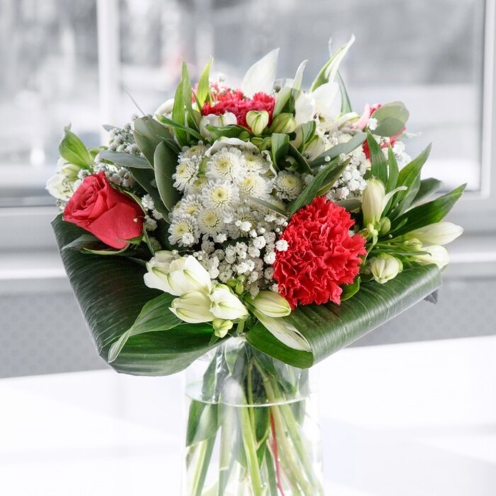 A finely balanced red and white floral arrangement stands centre stage in a clear cylindrical glass vase, the stems below the surface arranged in clean, parallel lines that speak of skilled hands and fresh stock. At the forefront, a ruffled crimson carnation bursts with intricate folds and texture, echoed by the lush, velvety depth of several deep red roses whose petals spiral outward in perfect, romantic curls. Threaded between these passionate reds are serene white blooms: alstroemerias with slim petals and faint speckling, rounded white chrysanthemums, and several tight white lily buds held close, hinting at elegant flowers still waiting to unfold over the coming days. A sprinkling of tiny white gypsophila, or baby's breath, drifts lightly through the bouquet like a dusting of first snow, giving the arrangement an airy softness and a sense of movement. The entire composition is wrapped and supported by an array of greenery-broad, dark green leaves polished to a gentle shine and carefully curled to form a sleek collar around the base, complemented by varied green foliage reminiscent of eucalyptus and slender, pointed leaves that add height, texture, and visual rhythm. The bouquet rests on a smooth white tabletop, set against a softly blurred, luminous background that could be a bright Brent Cross living room or an office near Prince Charles Drive, washed in daylight. The lighting is calm and even, enhancing every colour and contour without glare, making the arrangement feel both modern and timeless. It is easy to imagine this bouquet as a centrepiece at a family celebration, a thank-you gift delivered across Brent Cross by a trusted local florist, or a thoughtful gesture to bring warmth and beauty into someone's everyday space.