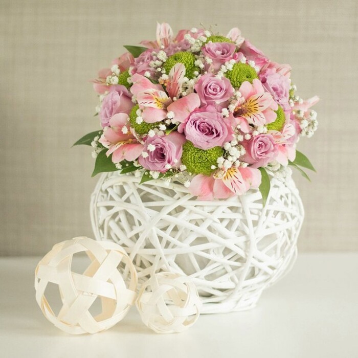 A striking floral centerpiece welcomes the eye from a sun-drenched Brent Cross table, its abundant pastel blooms a nod to both contemporary design and local charm. Central to the composition are plush, pastel pink and lavender roses, each unfurling their velvety layers in full bloom-a visual marvel reminiscent of the first blooms in Brent's neighborhood gardens. Pink alstroemeria, with their telling trumpet shapes and inner striped markings, nestle among the roses, offering rhythm and elegance. Contrasting the delicate pastels, clusters of lively lime green button chrysanthemums punctuate the display, their rounded heads vivid and unique. Throughout the bouquet, airy white sprays of baby's breath interlace, imparting an ethereal softness and enhancing the arrangement's sense of abundance. Glossy dark green leaves emerge between floral clusters, anchoring the entire piece with deep, natural tones reminiscent of leafy avenues stretching toward Brent Cross Shopping Centre. The arrangement is cradled in a statement white vase, its open, woven structure lending a contemporary yet handcrafted essence that echoes local North London craftsmanship. Flanking the composition, two decorative spheres-crafted from light, woven strands and sitting at staggered heights-add artistic symmetry, mirroring the vase's latticework. The neutral, textured beige background provides a calm canvas for the array of colour and design. This image effortlessly fuses freshness, joy, and local artistry, perfect as a Brent Cross wedding centerpiece or for elevating special gatherings with understated elegance.