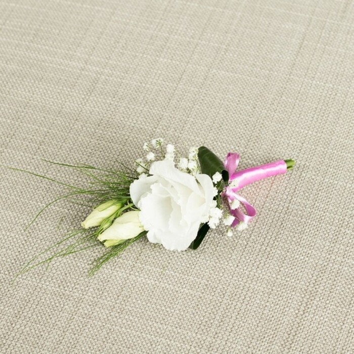 A finely detailed wedding boutonniere is captured from a gently elevated angle, resting at a diagonal on a warm, light beige fabric that mimics the texture of natural linen, its subtle weave adding depth and softness to the scene. At the heart of the arrangement is a single, fully bloomed white eustoma, or lisianthus, its many ruffled petals unfolding outward in delicate layers, creating a plush, rounded flower head that feels both contemporary and timeless. Just to the left and slightly beneath the main flower, two slender buds from the same eustoma stem stretch forward, their creamy, pale green forms still tightly closed, suggesting imminent bloom and the quiet sense of a day just beginning. Interspersed among these focal flowers are tiny clouds of white gypsophila, or baby's breath, their miniature blossoms forming an airy halo that softens the composition and lends it a romantic, dreamlike quality. Behind the flowers, slender, grass-like blades of foliage fan gracefully towards the lower left corner, their rich, fresh green tones contrasting beautifully with the white petals, while a single broader, darker green leaf sits discreetly behind the main bloom, giving structure and a sense of grounding. At the base, the stems are neatly aligned and bound together by a vivid fuchsia satin ribbon, tied into a neat, perky bow that introduces a joyful burst of colour. The satin's smooth sheen catches the soft, even lighting, making the pink appear almost luminous against the neutral backdrop. The overall impression is of a carefully considered floral accent, the kind a seasoned Brent Cross florist might pin onto a groom's lapel before a ceremony near Clitterhouse Playing Fields, capturing a balance of elegance, freshness, and a playful hint of personality.