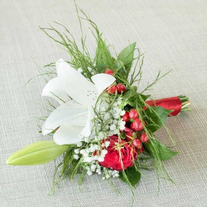 Resting delicately atop a soft, pale, textured surface evocative of local Brent Cross home interiors, this bridal bouquet presents a beautifully balanced palette and expert craftsmanship. At the focal point blooms a stunning white lily, petals wide and pure, decorated with gentle russet speckles near the centre and surrounding luminous golden anthers. Beneath and just aside, a lush red rose, velvety and rich, unfolds its tightly layered petals, echoing the romance of weddings held in Brent Cross's beloved venues. Between these vibrant cores, tiny white clouds of baby's breath scatter a gentle softness, reminiscent of northern spring mornings in the nearby parks. Shining clusters of bold red berries add a lively seasonal accent, recalling the energetic markets of the area. The arrangement is embraced by varied greenery, from wispy fern-like fronds to substantial, dark leaves that amplify both vibrancy and depth. A pale green lily bud peeks forth, promising future beauty and hope. This harmonious bouquet is tied at the base with a glowing red satin ribbon, the finish shimmering gently and unifying the bold reds found throughout. The composition, ideal for honouring bridesmaids, celebrating anniversaries, or sharing affection in Brent Cross, showcases the sensitivity and skill of local florists, creating a bouquet that resonates with love, festivity, and enduring charm.
