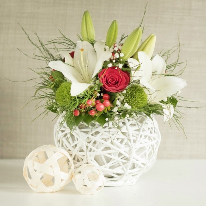 A beautifully composed floral centerpiece is shown resting on a sleek white tabletop, the kind you might find laid out for a wedding breakfast near Brent Cross Shopping Centre or an elegant celebration in a Hendon venue. The focal point of the image is a distinctive, globe-shaped vase in pure white, constructed from slender, overlapping strands that create an open, basket-weave sphere. The design allows light to slip through its gaps, casting soft shadows and adding a subtle sculptural quality to the arrangement. Filling this contemporary vessel is a lush bouquet that blends white lilies, red roses, green pompon blooms, and layers of fresh foliage. Several lilies have fully opened, their broad, creamy petals sweeping outward to frame golden stamens and faint speckling at the throat, evoking a calm, almost bridal purity. Upright green lily buds punctuate the top of the arrangement, promising new blooms and extending the life of the display. Amid the lilies, one particularly striking red rose claims the centre, its velvety petals coiled into a tight spiral, while additional roses in similar deep red tones support its impact and bring a romantic depth of colour. Rounded, pompon-like green dianthus or chrysanthemums introduce an unexpected twist, their soft, tufted texture and bright hue lifting the overall palette. Fine sprays of white baby's breath drift through the composition like delicate clouds, creating lightness, while discreet clusters of red berries echo the rose tones and lend a gently festive accent. Long, wispy strands of asparagus fern and darker green leaves spill outward from the vase, giving the arrangement a sense of organic flow and movement. To the left of the main display, two pale, woven decorative spheres rest quietly, echoing the vase's texture and completing a tranquil, sophisticated scene that suits both weddings and intimate gatherings across Brent Cross and the surrounding neighbourhoods.