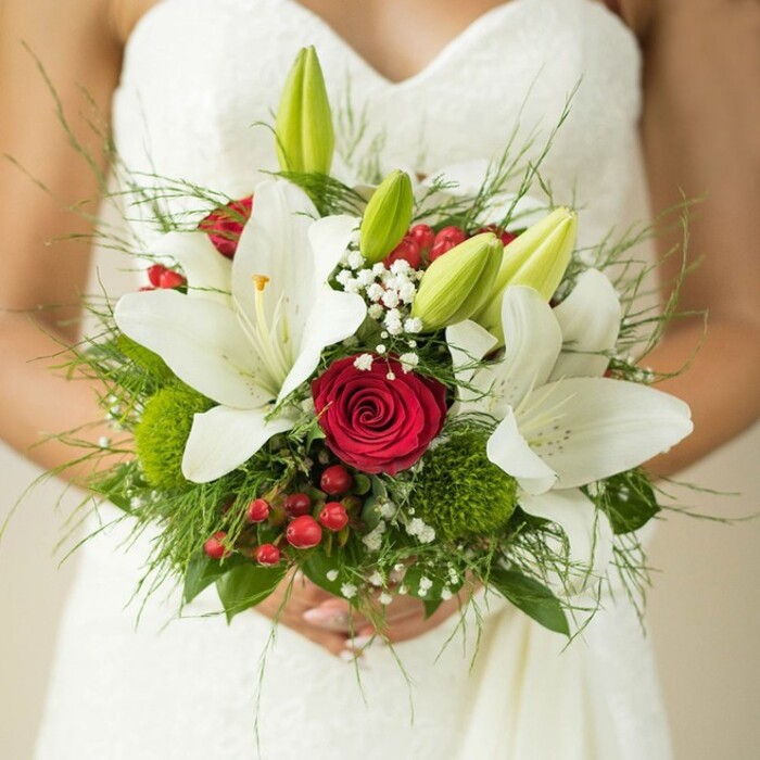 A detailed close-up reveals a bride's hands, softly clasped around an exuberant bridal bouquet-set against the luminous backdrop of her white dress, likely near Brent Cross's lively avenues or a local ceremony hall. The bouquet is a celebration of colour and form: large, snowy white lilies, their blooming faces tipped with golden accents, evoke a sense of timeless grace. Emerging green buds promise the arrival of new chapters, while a perfectly placed, rich red rose commands the centre, its furled petals embodying true romance. A constellation of white gypsophila sparkles among spheres of green trick dianthus and scarlet hypericum berries, producing a kaleidoscope of textures as light dances across the arrangement. Lush greenery, uniquely gathered from florists familiar with Hendon or Golders Green, nestles at the base, combining ferns and wide leaves for fullness. The entire ensemble reflects both tradition and spirited celebration, ideal for brides seeking to mark their vows with a locally inspired floral masterpiece-whether it be at a Brent Cross venue or garden ceremony.