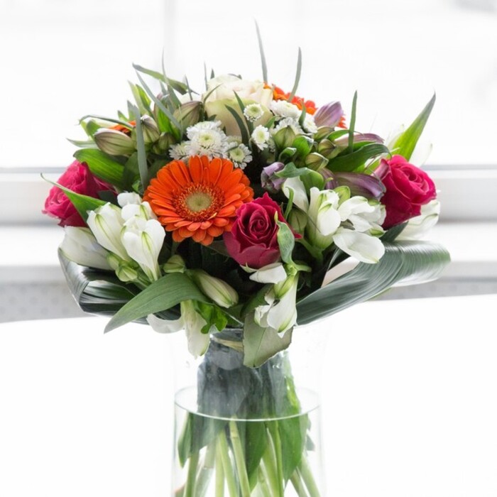 This sumptuous bouquet, conceived by expert florists in Brent Cross, is a celebration of balanced colour, robust structure, and heartfelt expression. Creamy gerberas form a luminous ring around brilliant orange hearts, their petals soft and inviting, recalling festive occasions in the heart of Cricklewood Lane. The deeply saturated fuchsia roses bring a touch of opulence, their tightly coiled petals suggesting enduring romance and sophisticated beauty. Ivory alstroemeria and petite white spray roses lace through the ensemble, imparting freshness and a light, garden-inspired touch reminiscent of breezes through Claremont Park. Abundant deep green foliage provides a sturdy nest for the blooms, with broad aspidistra leaves artfully encircling the base, strengthening the arrangement's rounded silhouette. Between these, slender greens and sprigs of fern lend organic movement and depth. Nestled in a crystal-clear, slender vase, the stems stand testament to freshness and skilled arrangement. Softly diffused daylight enhances each flower's colour, avoiding harshness and maintaining a tranquil, rejuvenating mood. The subtle, imagined fragrance-a mix of lilies and green leaves-lingers in the air, evoking memories of open spaces near Brent Cross. This bouquet feels tailor-made for celebratory brunches, heartfelt thank-yous, or simply bringing a vibrant streak of joy to a loved one's table. Each detail reflects the community spirit and expertise of Brent Cross's dedicated florists.