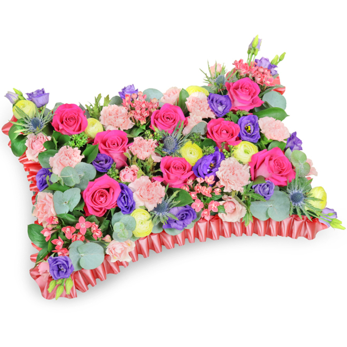 Captured from an oblique, slightly elevated viewpoint against a stark white background, this sumptuous pillow-style floral tribute presents a tapestry of colour and texture that feels both comforting and celebratory. The arrangement overflows with hot pink roses - some tightly coiled, others opening into soft velveteen cups - which are artfully contrasted by clouds of pale pink carnations that introduce a frilly, tactile dimension. Deep purple lisianthus provide bell-shaped accents, while bright lemon-yellow ranunculus and creamy small fillers inject sunlight into the composition. Intermittent sprays of silver-blue Eryngium add a cool, sculptural edge, their spiky forms giving rhythm amid the lush petals. Lush greenery, notably rounded eucalyptus leaves in their silvery-green tone, and darker filler foliage create layers of depth and a calming, herbal fragrance that complements the sweeter rose scent. The whole piece rests upon a ruffled, deep red satin ribbon base that frames the cushion with a refined, pleated border and reinforces the rectangular silhouette. Soft, directional light brings out the sheen of petals and ribbon, while the dense packing and concealed structural support give the cushion a reassuring, pillow-like presence. Suited to funerals, memorial services or placing at a graveside, this floral pillow communicates warmth, remembrance and the celebration of a cherished life - a sentiment well suited to families throughout Brent Cross, near the shopping centre and neighbouring streets, seeking a sensitive, handmade tribute.