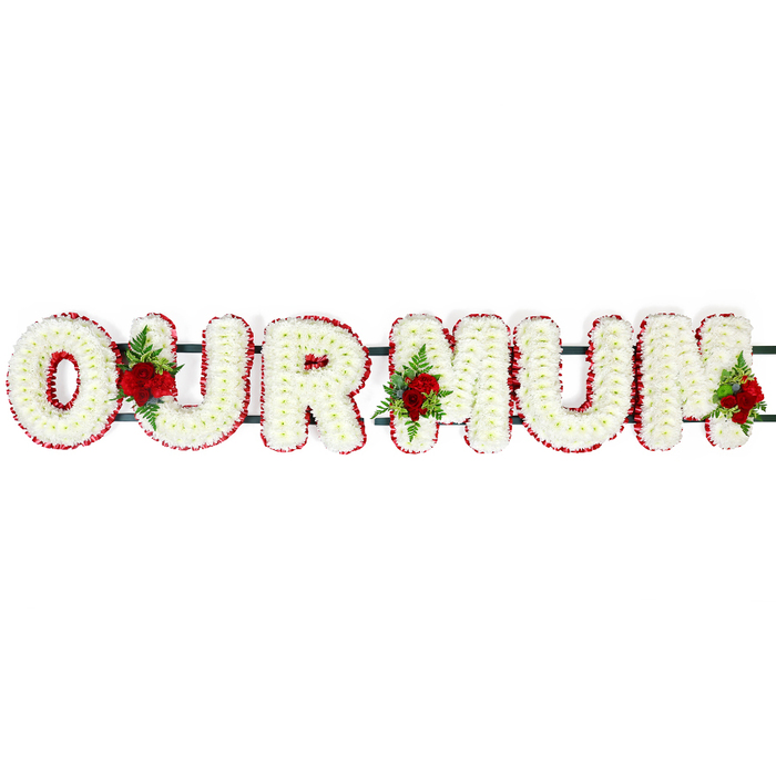A tender 'OUR MUM' floral letter tribute is laid out in a straight horizontal line, photographed against a simple white background so that the focus remains solely on the craftsmanship of the flowers. Each bold capital letter is densely packed with pure white chrysanthemum blooms, their compact petals forming a smooth, domed texture that looks almost like soft quilting made from petals. The chrysanthemums create a consistent, snowy surface across the whole word, suggesting peace and purity, like a calm pause before family and friends gather in Brent Cross to pay their respects. Around each letter runs a distinct border of deep red flowers, probably carnations or other tight-headed blooms. These red petals are clustered closely together, building a rich, velvety frame that outlines the contours of every letter with clarity, giving the tribute definition and visual strength. The contrast between the crisp white centres and the bold red edges is striking yet harmonious, echoing both the gentleness and the deep emotion felt when remembering a beloved mother. Nestled between sections of the lettering are three sprays of deep red roses, each arranged with lush green foliage and delicate fern. One spray sits securely in the gap between the O and U, another lies between the U and the first M, and the final grouping softens the outer side of the last M, as though gently closing the phrase with a final embrace of roses. The rose heads are slightly open, their layered petals giving a sense of depth and movement. Behind the flowers, subtle hints of dark green framework can be glimpsed, connecting and supporting the letters so the tribute can be placed on a coffin, hearse, or display stand. Altogether, the piece feels both stately and intimate, a traditional yet personal arrangement that speaks of love, gratitude, and respectful farewell to a cherished mum.