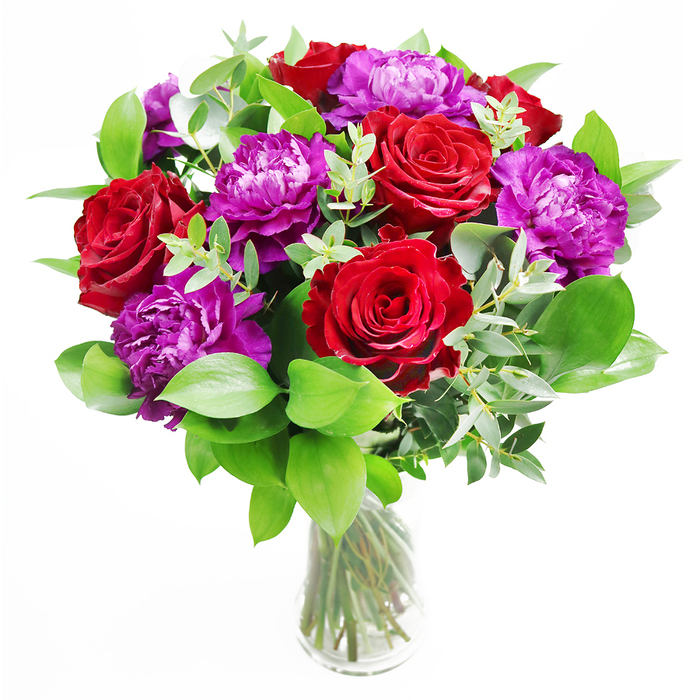 In this floral arrangement, classic romance meets modern elegance, with deep red roses and sumptuous purple peonies gathered together in a clear glass vase, much like a showpiece you might place in a Brent Cross living room after a special delivery. The roses stand proudly at the heart of the bouquet, each bloom generously open, revealing layers of velvety petals in rich crimson tones that feel both passionate and reassuringly timeless. Woven between them, the peonies provide a softer, more billowing presence: their blossoms are large and rounded, formed from a multitude of ruffled petals in a sophisticated purple that shifts from gentle to intense depending on how the light touches them. Surrounding this central cluster of blooms is a halo of greenery, adding structure and freshness. Wide, glossy leaves fan out and curve slightly, giving the bouquet a sense of fullness, while trails of eucalyptus snake gently through the design, their slim stems and muted silver-green leaves lending airiness and a touch of refinement. The glass vase is intentionally understated, its transparency showcasing the neatly arranged stems and the clarity of the water, almost as if the bouquet has just been placed after being hand-delivered from a nearby Brent Cross florist. Light falls across the whole composition, creating delicate highlights on the petals and soft shadows in the folds and overlaps of the leaves. You can almost imagine the scent lingering in the room: a romantic rose fragrance punctuated by the fresh, cool note of eucalyptus. This bouquet feels ideal for an anniversary dinner near the Brent Cross Shopping Centre, a grand gesture of admiration, or as a striking arrangement to celebrate a significant life moment with style and warmth.
