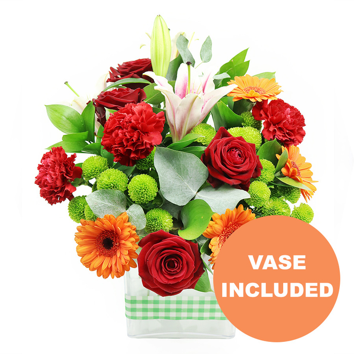 A generous bouquet of fresh flowers is shown in this image, its colours and textures carefully orchestrated like a mini garden placed in a glass vase, perfect for a home or office near Brent Cross. The flowers are arranged in a low, rectangular clear glass container, the water-filled interior and clean edges giving a smart, contemporary appearance. Wrapped horizontally around the bottom half is a light green and white gingham ribbon, adding a soft, domestic touch that evokes relaxed weekend gatherings after a shopping trip or stroll through local parks. At the heart of the bouquet, deep crimson red roses stand in full, velvety clusters, each bloom composed of tightly layered petals that create a sense of depth and luxury. Around these, red-orange carnations with delicately frilled edges add a sense of movement and intricate texture, softening the arrangement's overall structure. Bright orange gerbera daisies are dotted throughout, their flat discs of petals arranged around dark central eyes, drawing attention with their sunny, extroverted character. Rising above the main cluster of flowers are white Oriental lilies, some fully opened into star-shaped blossoms streaked lightly with pink along the inner petals, others still in pointed buds. These lilies give the bouquet height and elegance, reminiscent of tall trees glimpsed beyond the rooftops near Brent Cross. Nestled between the larger blooms are small tufts of lime-green pompom chrysanthemums, perfectly rounded and vivid, creating delightful pops of fresh colour and adding a playful, almost bubbly texture. Encircling the arrangement, lush foliage in varying greens-broad, glossy leaves mixed with slender, silver-green eucalyptus stems-creates depth, structure, and a slightly wild, garden-inspired feel, as though this arrangement has just been composed by Brent Cross Flowers for same-day delivery. The clean white background ensures every petal and leaf stands out clearly. In the bottom right corner lies a circular orange overlay, partly overlapping the vase, printed with VASE INCLUDED in bold white capitals, indicating that the pictured container is included with the bouquet.
