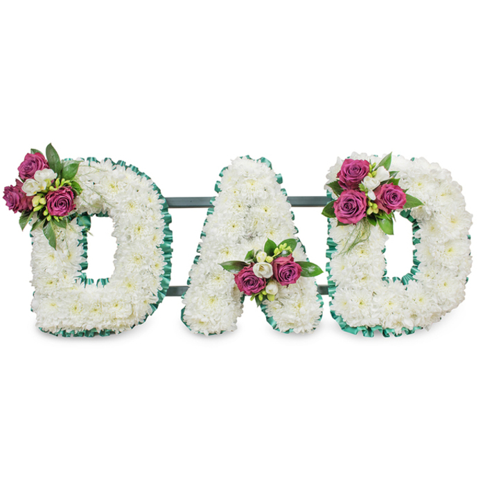 A carefully composed memorial tribute spells DAD in three substantial, three-dimensional letters, each letter densely covered in pristine white chrysanthemums that give a soft, cloud-like surface and a sense of quiet purity. The letters are sharply defined at their edges by an emerald green, ruffled ribbon that outlines both the outside and the internal counters, lending a touch of formality while keeping the feel tender. A muted dark green-grey connecting bar sits just behind the letters, suggesting the kind of solid support used for chapel or graveside displays. Rich magenta roses punctuate the arrangement in three discreet clusters - upper left of the first D, nested in the lower V of the A, and upper right of the final D - each cluster threaded with delicate white freesia and lush deep-green foliage to enhance contrast and depth. The interplay of textures is notable: the soft chrysanthemum cushions, the velvet of the roses, and the slender, fragrant freesia stems combine to create a multi-sensory tribute whose subtle scent and visual restraint feel appropriate for remembrance. Shot against a clean white background, the composition reads with dignity and warmth. Brent Cross Flowers crafts this style of bespoke memorial lettering for services across Brent Cross, NW11, and nearby Golders Green and Hendon, offering an arrangement that communicates affection, honour, and a calm farewell for a beloved father.