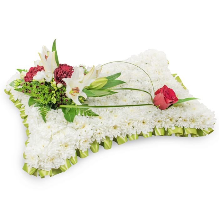 A classic funeral cushion, photographed against a pure white backdrop, that blends traditional formality with delicate craftsmanship for moments of remembrance in Brent Cross and beyond. The rectangular base is densely covered with hundreds of small white chrysanthemums, each bloom positioned to create a sumptuous, seamless surface that suggests softness and repose. A pale lime-green satin ribbon, pleated into a neat frill, encircles the whole pillow and adds a whisper of colour and refined finish against the white mass. Anchoring the design on the upper left is a richly textured cluster: two pristine white Oriental lilies, fully open with yellow stamens visible, stand beside a closed lily bud; nearby, deep burgundy spray carnations provide weight and contrast, their ruffled petals introducing darker, comforting tones. An assortment of green foliage-ranging from feathery fern fronds to broader glossy leaves-forms a verdant backdrop for the blooms, giving depth and natural movement. From that cluster a graceful arch of bright green salal or decorative wire sweeps across the chrysanthemum field toward the right, terminating in a single deep red rosebud poised to unfurl, a poignant emblem of enduring love and respect. Thoughtfully composed and suitable for placement at chapel services near Brent Cross Shopping Centre, at family homes in Cricklewood or at cemeteries around Golders Green, this sympathy pillow embodies purity, peace and heartfelt condolence. Prepared by experienced hands at Brent Cross Flowers, it balances texture, line and colour to offer comfort and a dignified tribute for grieving families.