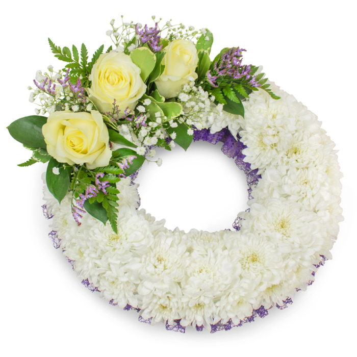 A dignified circular wreath, photographed on a plain white background and intended as a sympathy or funeral tribute for families across Brent Cross, Golders Green and nearby Hendon. The foundational ring is composed of hundreds of small white chrysanthemums, each compact head creating a soft, cushiony texture that reads as calm and reverent from a distance and up close. A delicate, dark purple mesh or ribbon peeks through the chrysanthemum mass along both inner and outer rims, introducing a subtle tonal frame that deepens the otherwise pristine white surface. At the upper-left quadrant a considered spray of accents provides the emotional focal point: three creamy yellow roses with unfurling, velvety petals suggest warmth and remembrance, while fine sprays of baby's breath lend an airy, cloud-like quality around them. Small lavender-purple statice (or similar filler blooms) add restrained pops of colour that harmonise with the purple trim. The greenery-mixtures of glossy broad leaves and lighter, fern-like fronds-gives structural support and a fresh, living contrast to the pale blooms. The even, soft lighting casts a gentle shadow beneath the wreath, giving it dimension and a sense of presence suitable for laying near a coffin or on an easel at a memorial service. As a local florist I assemble pieces like this to express condolences with careful craftsmanship and sensitivity; Brent Cross Flowers can prepare and deliver this traditional wreath for a family service within the local area, offering a respectful, composed expression of sympathy and remembrance.