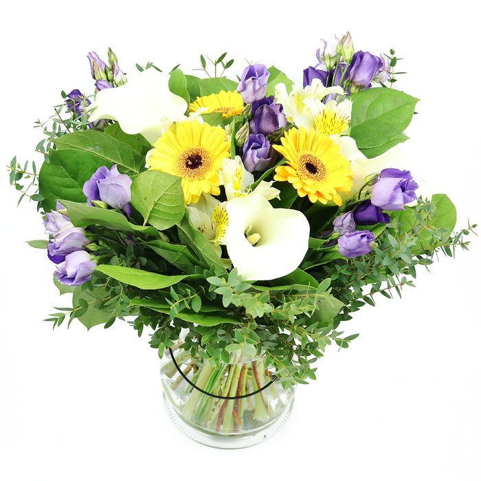 An opulent interpretation of a mixed floral bouquet by Brent Cross Flowers, poised in a crystal-clear glass vase against a pristine white background. The composition glows with pure sunshine from large yellow Gerbera daisies, their daisy-bright centers forming bold anchors throughout the arrangement. Nestled amongst them are pure white Calla lilies with flawless, trumpet-shaped petals, exuding a refined elegance that elevates the bouquet's luxury aura. Lavender to rich purple Lisianthus provide sumptuous depth and graceful curves, some blossoms fully unfurled to reveal their velvet petals, others remaining as artful buds. Accents of small white Alstroemeria weave subtle yellow and green streaks, adding delicate texture. The verdant foundation is abundant, featuring eucalyptus branches with their silvery sheen and a mix of broad, deep green leaves that lend structure and a lush, fashionable fullness. The glass vase reveals neatly trimmed stems resting in crystal-clear water, while a slender black ribbon secures the look with a touch of contemporary polish. This refined arrangement conveys celebration, appreciation, and refined taste, perfect as a centerpiece for stylish Brent Cross events or for gifting someone special in Hendon or Cricklewood with same-day delivery from Brent Cross Flowers.