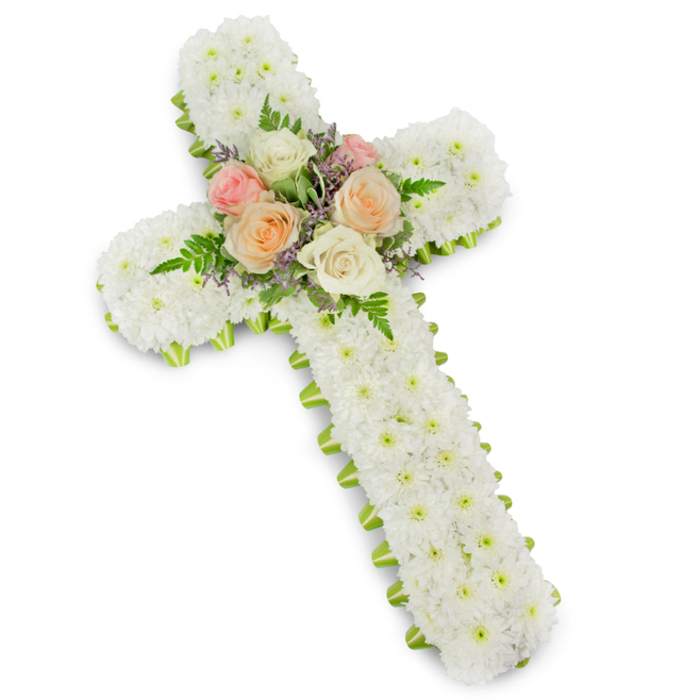 In this photograph, a traditional funeral flower cross is presented against a brilliant white background, the clarity of the setting enhancing every nuance of the design. The cross is robust and well-defined, formed entirely from compact clusters of white chrysanthemum button blooms that cover the frame without any visible gaps. The result is a remarkably even, velvety texture that suggests softness to the touch, as if one could press a hand into the flowers and feel only petals. This unbroken white surface communicates innocence, serenity, and the quiet reassurance often sought during services in Brent Cross churches or memorial halls. At the precise centre of the cross, a rounded cluster of roses provides a gentle focal point. Several fully open roses, their petals unfurling in harmonious spirals, are arranged in an intimate gathering of hues: soft blush pink, subdued peach-apricot, and creamy ivory. Surrounding and threading between these roses are slender fern fronds, their deep green tips adding lift and movement, along with fine sprays of lavender or pale purple filler flowers that introduce a discreet secondary colour. Around the edge of the entire cross runs a narrow outline of lime green ribbon. It peeks out just enough to define the cross's silhouette and offer a fresh, contemporary accent without disturbing the overall calm. The lighting is soft yet precise, casting a gentle shadow that lends the arrangement dimension. This floral cross feels deeply reverent and compassionate, an ideal tribute for a Brent Cross family wishing to send a message of faith, hope, and lasting love to accompany a final farewell.