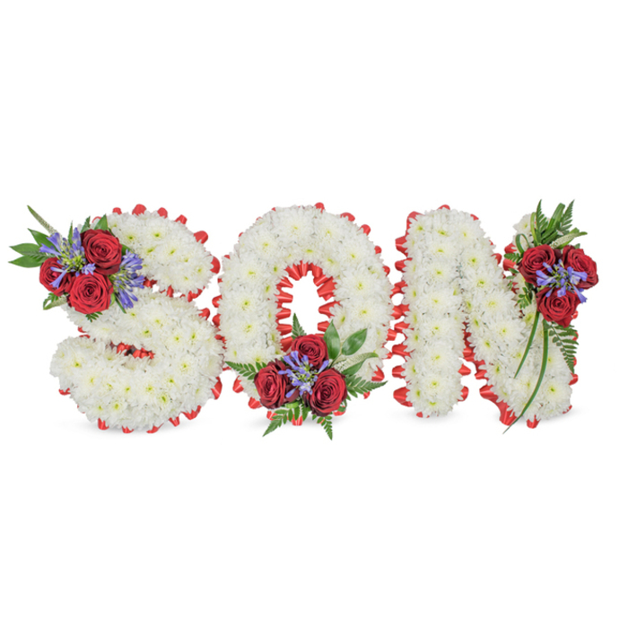 A meticulously crafted memorial arrangement spells the word SON in prominent three-dimensional letters, each letter densely constructed from small, immaculate white chrysanthemums that lend the surface a plush, cloud-like appearance. The letters sit against a crisp white background and are sharply defined by a folded band of bright red satin ribbon forming a scalloped border; the ribbon's sheen contrasts beautifully with the matte softness of the chrysanthemums. Each letter is embellished with a considered cluster of accent flowers and foliage: the S carries a composition of deep red roses and upright blue-purple spires at its upper left, the O is garnished with a compact spray at its bottom centre, and the N features a trailing cluster down its upper right stem. The blue-purple accents-Liatris or an equivalent spike-add verticality and a cool tonal counterpoint to the warm crimson roses, while the dark green ferns and leafy tendrils provide texture and a gentle trailing motion. The overall effect is dignified and tender: white symbolising peace and purity, red conveying love and remembrance. This bespoke floral lettering is ideal for a funeral, wake, or graveside service to honour a beloved son. As Brent Cross Flowers, serving Brent Cross and nearby streets around Staples Corner and the shopping centre, we design such tributes with careful attention to proportion, scent, and the quiet emotion families seek to express when saying goodbye.