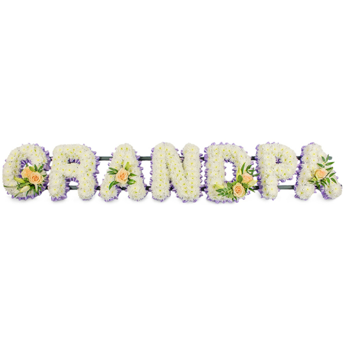 Set against a bright white background is a horizontal floral name tribute that reads GRANDPA, crafted with the kind of care and precision associated with experienced Brent Cross florists. Each uppercase letter stands clearly defined, yet the underlying structure is almost entirely hidden beneath a lush layer of flowers. The main body of each letter is made up of countless white spray chrysanthemums or similar small-headed blooms, nestled tightly together so that their soft petals form a continuous, pillow-like surface. You can imagine the faint, fresh scent of newly cut stems, the sort of fragrance that might greet you when you walk into a local flower shop just off the North Circular. Surrounding the white centres of every letter is a narrow edging of light lavender or lilac blossoms, creating a delicate frame that accentuates the typography and adds a gentle wash of colour. Along the sequence of letters, carefully placed accents of peach roses break the uniformity with warm, velvety spirals of apricot and cream. The G and D host slightly larger groupings, where the roses are interwoven with slender white lily buds and vibrant green foliage, spilling just beyond the edges and lending a natural, garden-like feel. The first A and the final A carry single peach roses, accompanied by small tufts of greenery that echo the fuller clusters without overwhelming the design. A thin dark bar running along the base indicates the support frame that allows the tribute to be displayed upright at a funeral or memorial service. The overall effect is elegantly understated yet deeply personal, perfectly suited for commemorating a much-loved grandfather from the Brent Cross area, perhaps after a lifetime of family gatherings nearby.