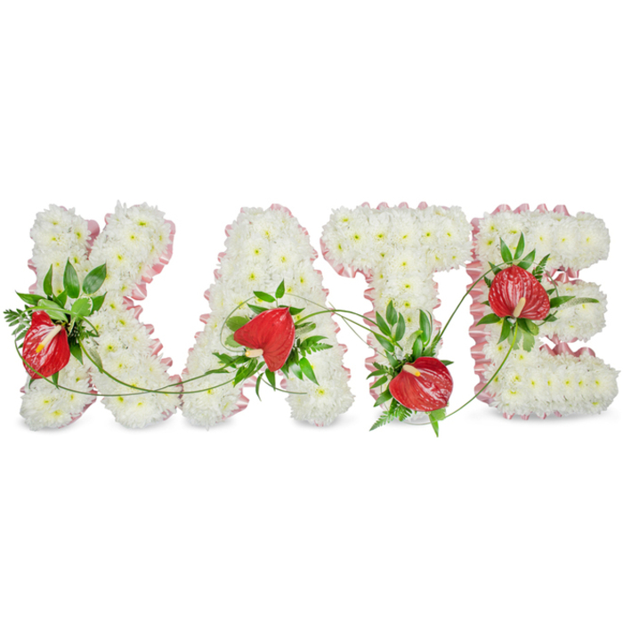 A carefully composed floral name tribute spells out KATE in large capital forms, each letter built from an even, dense carpet of small white chrysanthemums that present a serene, velvety texture. A pale pink, ruffled ribbon trims the outer edges of every letter, the pleats catching light and adding a soft, almost fabric-like frame to the floral sculpture. Bright red anthuriums are arranged as intentional focal points within the white fields-their glossy, heart-shaped spathes and prominent spadices providing a warm, vivid contrast that reads as affection within the solemn palette. Interspersed throughout the letters are deep green foliage elements-fern fronds, broad glossy leaves and sprigs of ruscus-introduced to bring depth, shadow and a natural, living quality to the otherwise uniform white surface. Thin green vines drift between the K, A, T and E, physically linking the names and suggesting a continuous line of remembrance. The display rests against a clean white background and casts a delicate drop shadow that accentuates the three-dimensional craftsmanship. As a florist who has prepared many tributes for chapels and local crematoria around Brent Cross Shopping Centre, I note the small details: the way the chrysanthemums are trimmed to a uniform height, how the ribbon edge is hand-pleated, and how each anthurium is angled to show its glossy face. The overall mood is peaceful and respectful-a composed, bespoke sympathy arrangement intended to honour memory with understated warmth and expert floral technique. Brent Cross Flowers crafts pieces like this for families across Brent Cross, Cricklewood and nearby neighbourhoods.