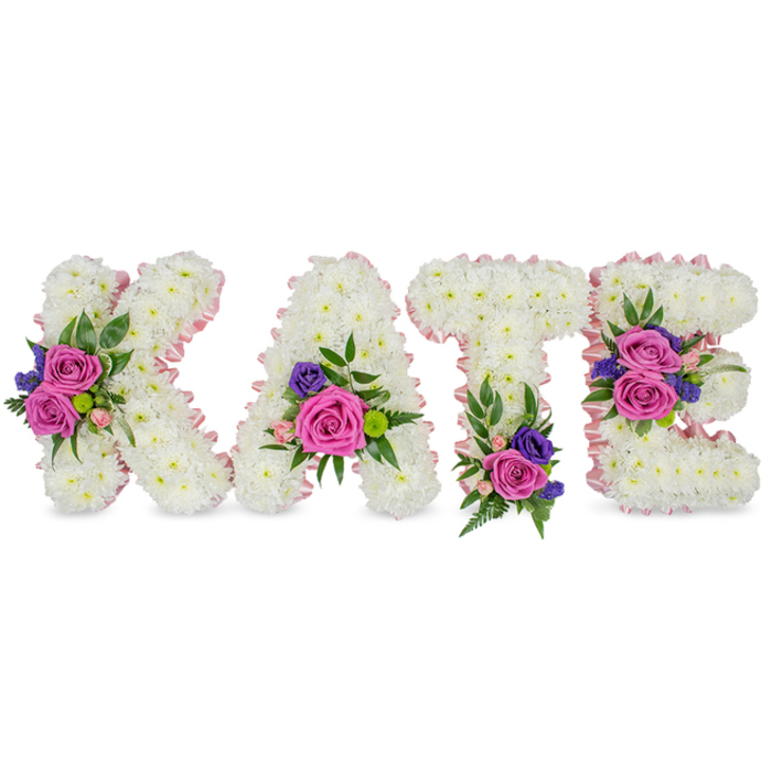 An expertly assembled memorial tribute spells out KATE in four freestanding, three-dimensional letters, each letter formed from a dense, even carpet of pristine white chrysanthemums that read like a soft, compact blanket of petals. A narrow, pale pink satin ribbon delicately follows the outer and inner contours of each character, adding a subtle halo of blush that defines the shapes against a clean white background. At carefully chosen points, vibrant clusters enliven the white: rich fuchsia-pink roses provide the main emotive note, joined by smaller pastel pink spray roses and shaded accents of deep purple or blue florals-likely statice or compact lisianthus-for contrast and depth. Varied foliage-delicate fern fronds, smooth eucalyptus leaves and fine green fillers-frames these clusters and gives gentle movement and texture. The placement is intentional and thoughtful: the K bears a prominent cluster on its diagonal stroke, the A is accented at the lower right curve, the T carries a bloom near its base, and the E is balanced with two clusters on the middle bar and the bottom arm, ensuring the name remains legible and heartfelt from every angle. Photographed in soft, even light that highlights texture and the faint scent of rose over chrysanthemum, this tribute is the kind of bespoke sympathy piece we prepare locally for services around Brent Cross Shopping Centre, nearby Hendon Lane chapels, or memorials in Fryent Country Park. Brent Cross Flowers crafts such personalised letters with attention to proportion, colour psychology and the quiet dignity needed when flowers speak for loved ones.
