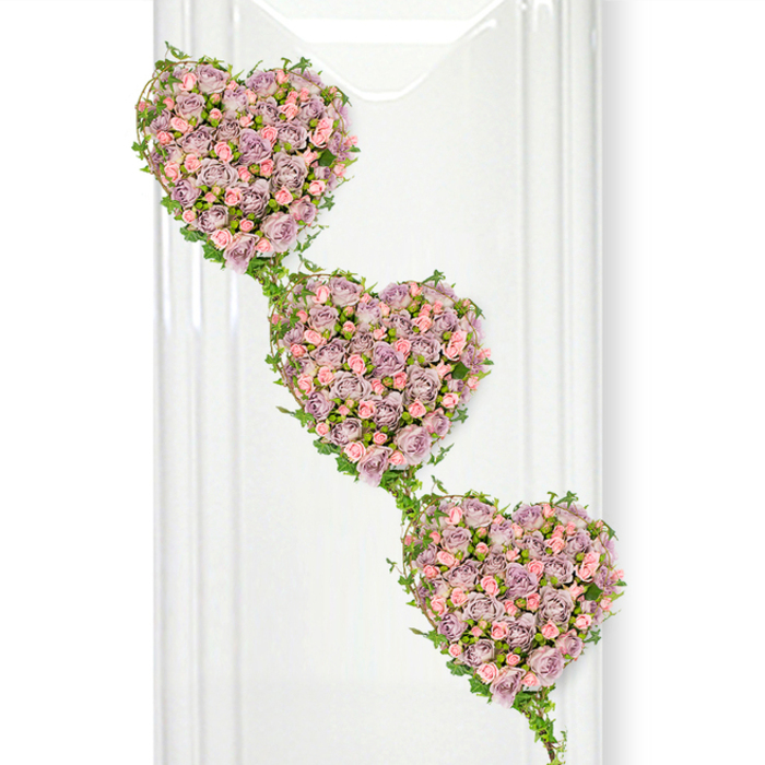 A trio of rose-covered hearts forms the centrepiece of this refined funeral tribute, captured in crisp detail against a pure white backdrop. The hearts are arranged in a flowing diagonal line, one beneath the other, as if carefully suspended in mid-air. Each heart is sculpted from dozens of individual roses, tightly packed so that almost no greenery shows through the full, velvety carpet of petals. The shades are gentle and romantic: dusty lavender, soft mauve, and pale shell-pink mingle seamlessly, with slight tonal variations that give the design a painterly quality. Some roses are still in bud, adding a sense of hope and tenderness, while others are wide open, their intricate centres visible, hinting at a life fully experienced. Encircling the shape of each heart, fresh ivy curls and loops, then reaches out to form the delicate connections between the three motifs. The ivy's glossy green leaves and sinuous stems add structure and a sense of natural movement, reminiscent of the climbing foliage that softens garden fences and pathways around Brent Cross and neighbouring suburbs. Gentle, diffused light washes over the flowers, eliminating harsh shadows and allowing each colour and texture to stand out. Subtle vertical reflections in the background suggest the presence of glass or polished surfaces, giving the scene a contemporary yet calm atmosphere, much like a modern chapel or quiet reflection room. The overall mood is one of tender remembrance and continuing love, making this triple-heart arrangement a thoughtful way to honour a loved one at a funeral, cremation, or memorial service, and a piece Brent Cross Flowers might create when a family wishes to express their grief and gratitude through something delicately beautiful.
