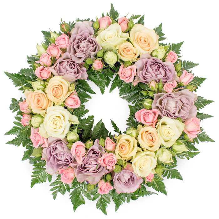 A beautifully detailed funeral wreath is pictured head-on against a spotless white backdrop, its circular form filled entirely with roses in pastel shades and a supporting layer of rich greenery. The wreath's ring appears lush and velvety, with no gaps, giving the impression of a continuous halo of petals. Dominating the arrangement are large, fully open roses in creamy white and soft ivory, their many overlapping petals curving outward to form generous rosettes that evoke purity and quiet devotion. Interwoven between these are roses in delicate pale peach and buttery yellow, introducing a gentle warmth reminiscent of the late afternoon light that falls across the streets around Brent Cross. Clusters of small blush pink spray roses flicker through the design, some blooms tightly furled, others just opening, adding complexity, texture, and a sense of unfolding memories. Dusty lavender and muted mauve roses are placed thoughtfully throughout the circle, their slightly smoky tones imparting a refined, antique feel that deepens the emotional resonance of the piece. Supporting all of this is a base of dark green fern fronds and similar fine-cut foliage. The feathery leaves project slightly beyond the line of roses, framing the circle and giving the wreath a soft, natural edge. The overall composition is symmetrical and perfectly round, symbolising continuity, eternal life, and lasting affection. It is the kind of carefully considered tribute Brent Cross Flowers might design for a funeral service at a local chapel or a memorial gathering held nearby, offering a quietly luxurious floral expression of sympathy, respect, and remembrance.