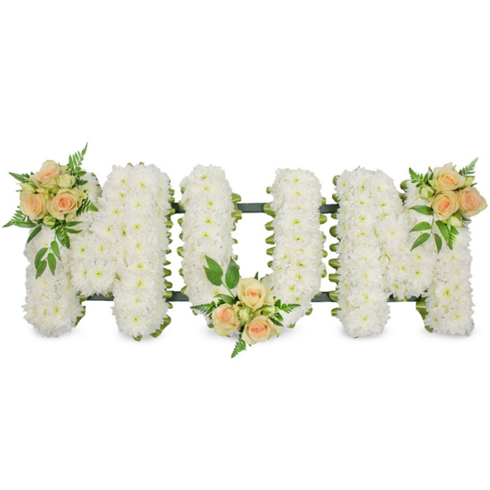 A carefully arranged floral tribute spells out MOM in large, elegant block letters, photographed against a spotless white background that accentuates the quality and care of the floristry. The three letters are mounted on an unseen supportive frame and generously padded with a dense covering of pure white flowers, likely small-headed chrysanthemums or spray carnations, each individual bloom pressed tightly beside the next. This creates a soft, quilted surface that is almost touchable, conveying an air of tranquility and respect well-suited to a funeral or memorial service in the Brent Cross area. A narrow band of dark green ribbon traces the outer edges of the letters, and the frame itself peeks through just enough to provide definition and structure, much as a local florist from Brent Cross Flowers would ensure for safe delivery to a nearby church or crematorium. Adding warmth and a sense of personal story, three rounded clusters of peach and apricot-coloured spray roses are placed strategically along the word. One rests near the upper left of the first M, one nestles into the lower central point of the O, and the final cluster is cradled at the upper right of the last M, drawing the eye gently from left to right. The roses themselves are at different stages of opening, with tight buds, half-open blooms, and lush, fully opened flowers all present, offering a subtle suggestion of different moments in a life. Each grouping is cushioned by sprigs of vibrant green foliage, from lacy fern-like leaves to stronger, glossy greens that add depth, contrast, and a touch of garden freshness. The overall arrangement feels both classic and personal, an eloquent floral message of love, remembrance, and gratitude for a mother, suitable for funeral services across Brent Cross, Hendon, and nearby Temple Fortune.