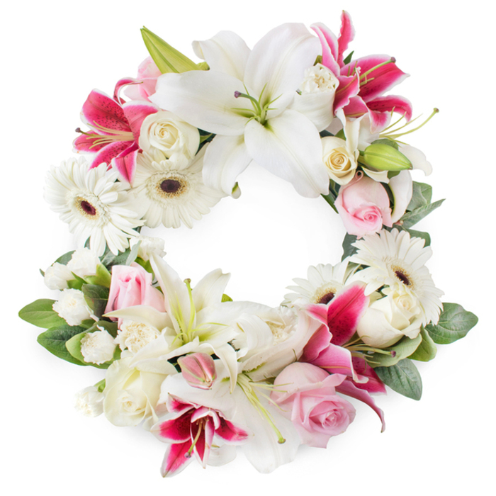Against a bright white backdrop, this circular funeral wreath appears like a serene floral halo, thoughtfully composed by an experienced local florist in the Brent Cross community. The ring is rich with large white Oriental lilies, whose elegant petals open wide to reveal green pistils and delicate golden stamens. These lilies bring a sense of purity and calm, their clean white tones speaking of peace and spiritual rest. Woven between them are deep magenta and fuchsia lilies, star-shaped and subtly speckled, adding vibrant splashes of colour that give the wreath depth and visual rhythm, much like the interplay of light and shadow along the paths near Brent Cross's shopping and residential areas. Throughout the design, clusters of soft pink roses and creamy white roses sit snugly among the larger blooms. Some roses remain as tight buds, symbolising life cut short or memories still unfolding, while others are fully open, their many layers suggesting a life well lived and the richness of shared experiences. Crisp white gerbera daisies with dark centres punctuate the wreath, their graphic shape lending a contemporary edge and a feeling of optimism. Smaller white filler flowers, probably miniature carnations or spray chrysanthemums, fill in every remaining space, creating a thick, plush texture that makes the wreath look abundant from any angle. The entire composition is encircled by lush green foliage, with leaves in deep and mid-green tones that frame the flowers and define the circular form. The result is a dignified, heartfelt sympathy wreath, perfect for funerals, memorials, or celebrations of life in Brent Cross and nearby North London, where families seek a floral tribute that blends classical elegance with gentle, modern touches.
