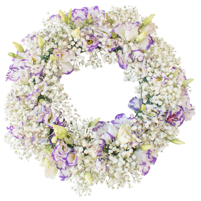 A round floral wreath lies against a bright, neutral white backdrop, where its refined mix of textures and tones stands out clearly. The foundation of the wreath is a generous ring of baby's breath, so heavily clustered that the individual Gypsophila stems merge into a thick, white cushion. Every tiny bloom adds to the illusion of a soft, snowy halo, creating a sense of lightness despite the fullness of the design. Interspersed across this white field are lisianthus blooms placed with practiced care, each one slightly different in its stage of opening. The lisianthus petals are soft and ruffled, with a base of creamy white that gently transitions into edges and veins tinted in lavender and deeper purple hues. A few flowers present fully unfurled faces, their layered petals spiralling around more shadowed centres; others are semi-closed or still neat in bud form, tipped with fresh green and faint yellow. This mixture of open flowers and buds gives the wreath a subtle sense of life and movement, as though it might continue to change over the coming days. There are no bold colours or extra ribbons; instead, the design leans on restraint and texture to convey its message. The open circle at the centre reflects themes of eternity and unending love, making the wreath a meaningful tribute for bereavements in the Brent Cross community. It is easy to imagine this arrangement being delivered to a quiet chapel, a crematorium service, or a family home, offering a gentle presence of beauty and solace, like a calm pause amid the bustle of the North Circular nearby.